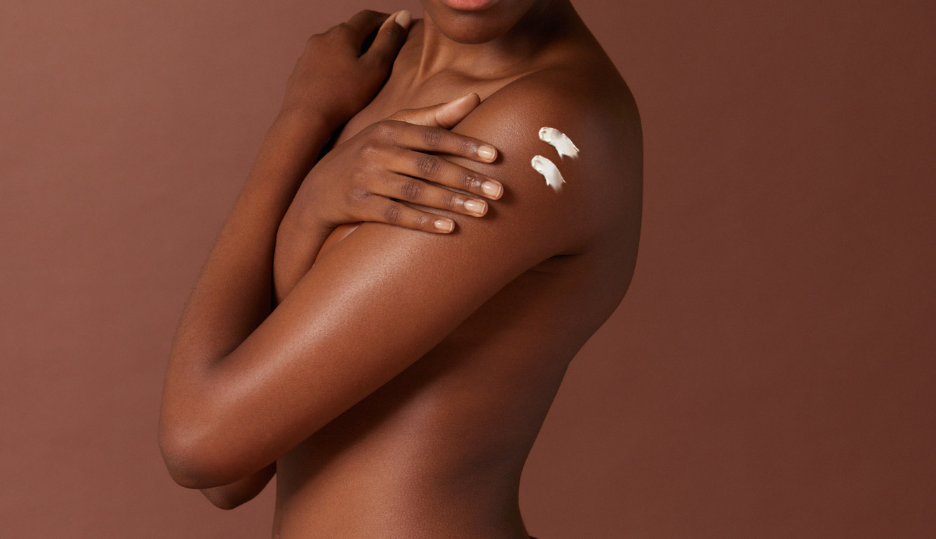 Close-up van een persoon met een donkere huid die zichzelf omarmt. Op de schouder zijn twee streepjes witte bodycrème aangebracht.
